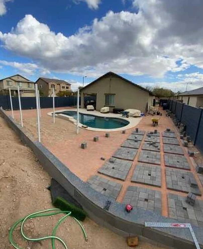 Paver installation in progress around a curved swimming pool with laid-out square paver sections.